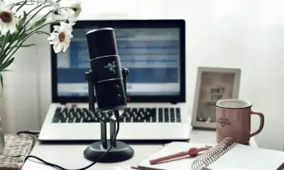 Microfone condensador preto para gravação de áudio com computador portátil ao fundo.