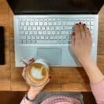 Teclado de laptop branco com uma mão segurando uma xícara de café e a outra teclado.
