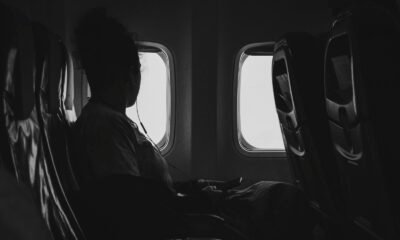 Imagem de uma passageira sentada em um avião, assistindo a um dispositivo eletrônico, com janelas ao.