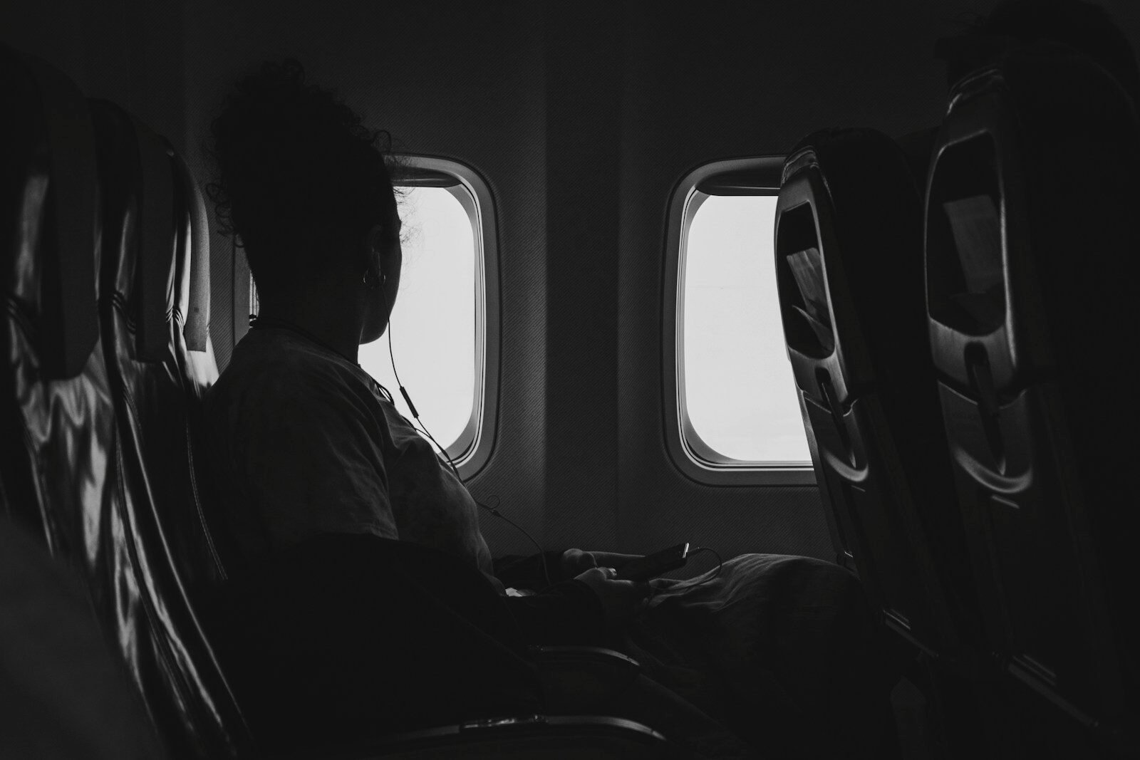 Imagem de uma passageira sentada em um avião, assistindo a um dispositivo eletrônico, com janelas ao.