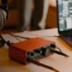Interface de áudio vermelha com controles, fones de ouvido e laptop com software de edição musica.